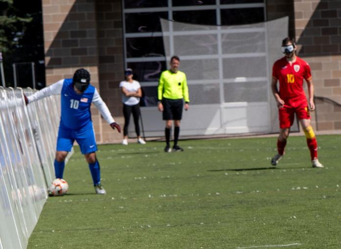 A blind football player wearing a blue number 10 jersey and an eye shade controls a soccer ball along the side barrier of a field. Another blindfolded player in a red number 10 jersey stands nearby